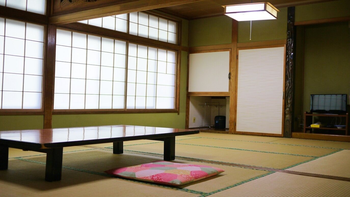 Ein Raum mit Tatami-Matten, einem niedrigen Tisch, Kissen, großen Shoji-Schirmen und einem Fernseher auf einem Ständer.