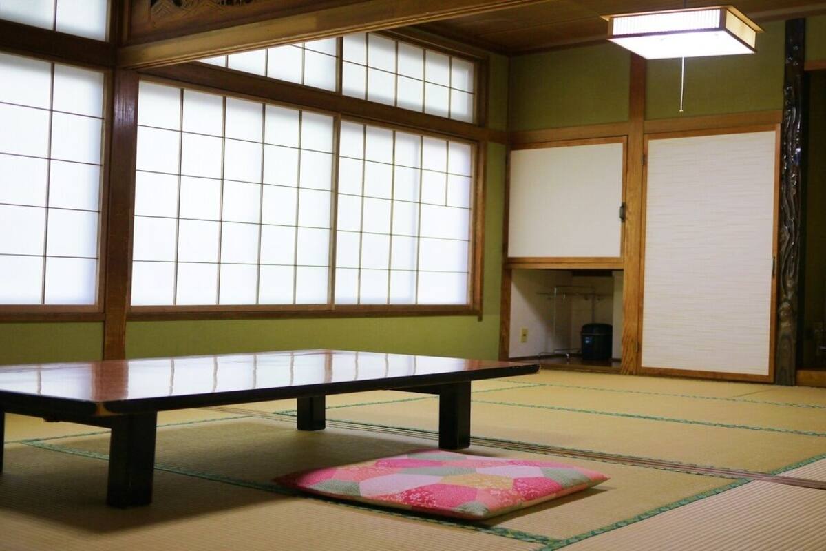 Une pièce avec un sol recouvert de tatamis, une petite table basse et des coussins, de grandes cloisons coulissantes en papier de riz (shoji) et un téléviseur posé sur un meuble.