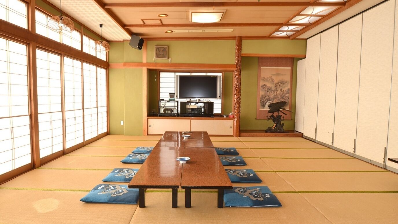 Ein Raum mit Tatami-Matten, einem langen Holztisch, Tatami-Matten, einem Fernseher und traditioneller Einrichtung.