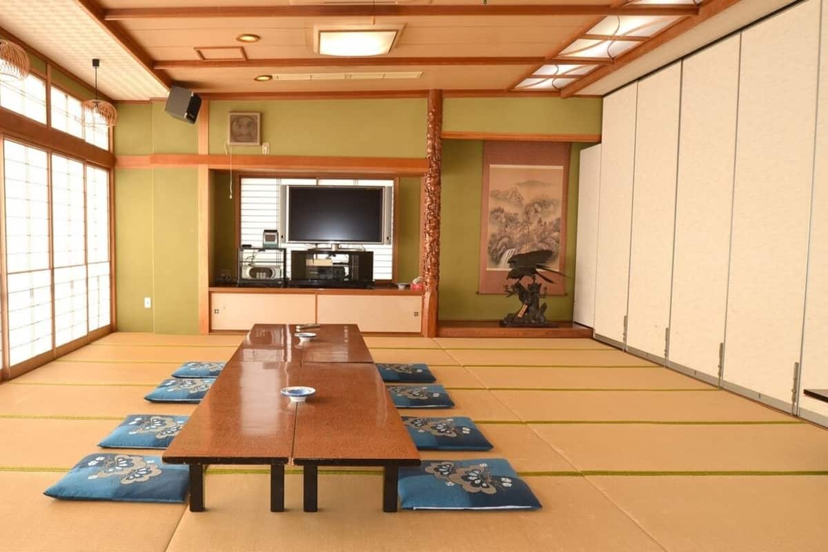 Une pièce avec un sol recouvert de tatamis, une longue table en bois, des tatamis, un téléviseur et un décor traditionnel.