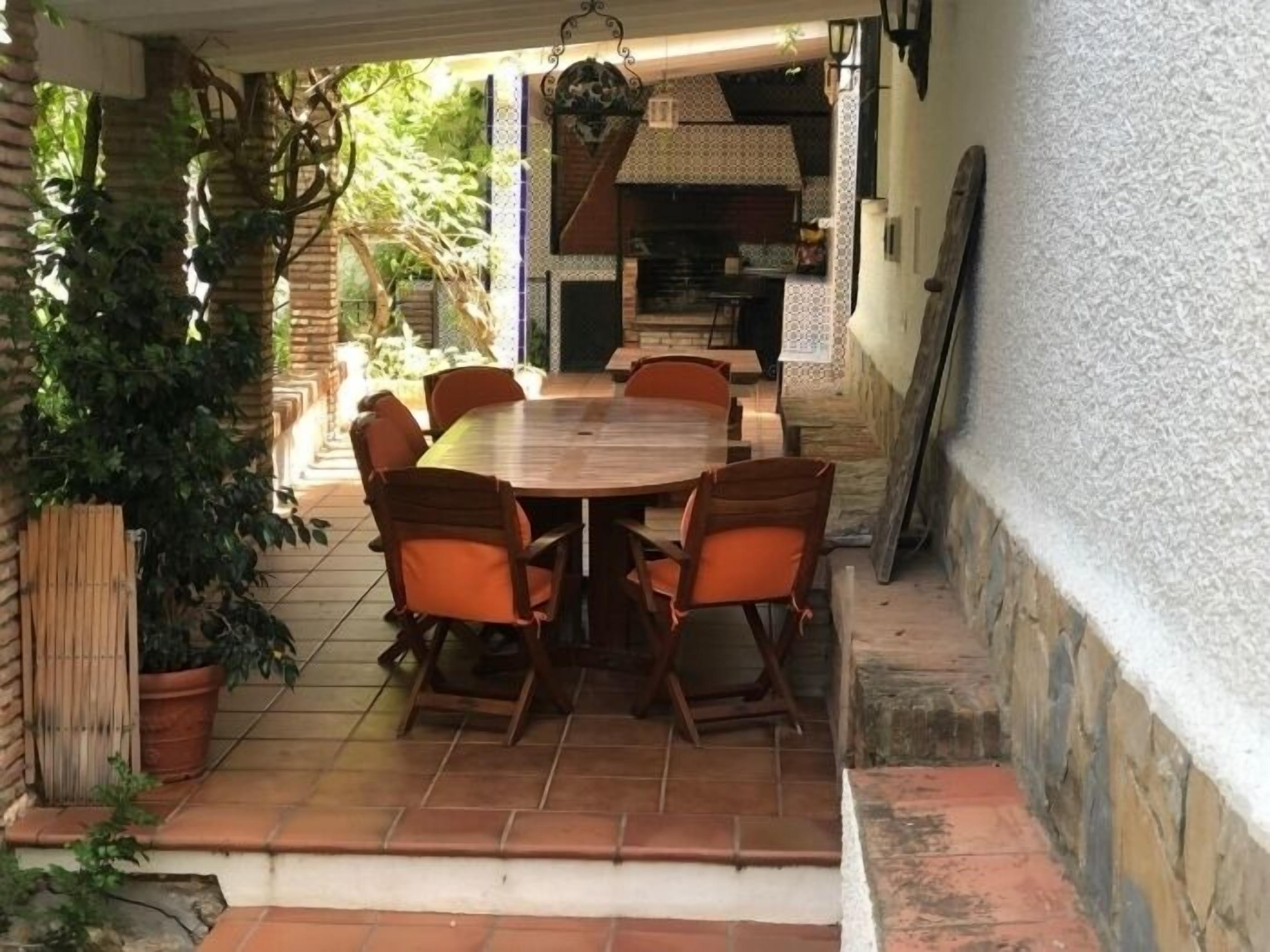 Une terrasse extérieure avec une table et des chaises en bois, entourée de plantes et d’un mur en briques.