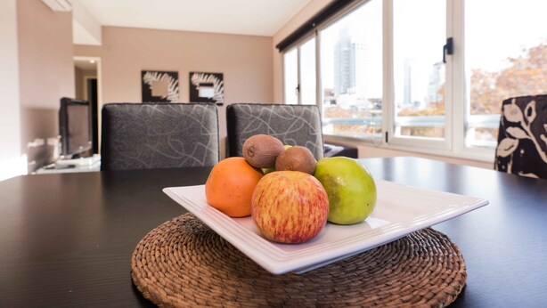 A dining table with a woven placemat, a white plate with fruits, and a city view through large windows.