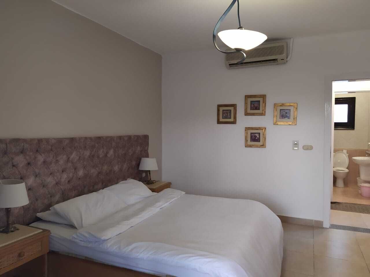 A hotel room with a large bed, two bedside tables with lamps, a ceiling light, and a wall-mounted air conditioner.