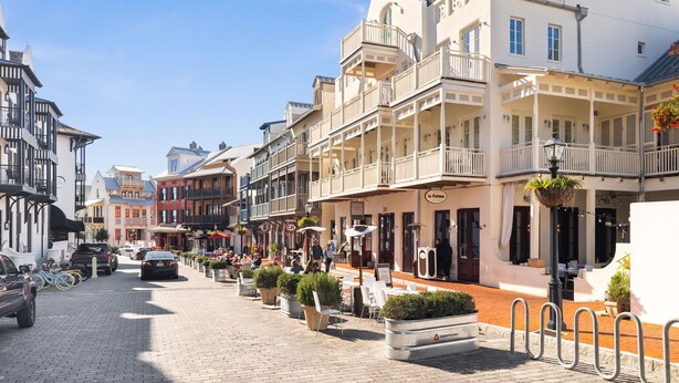 Une rue pavée bordée d’immeubles à plusieurs étages, de terrasses et de quelques voitures garées.