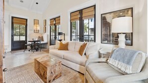 Un salon spacieux avec un grand canapé, une table basse et un fauteuil à rayures. De grandes fenêtres sont équipées de stores, et on y trouve une cheminée ainsi qu’un coin repas avec une table et des chaises.