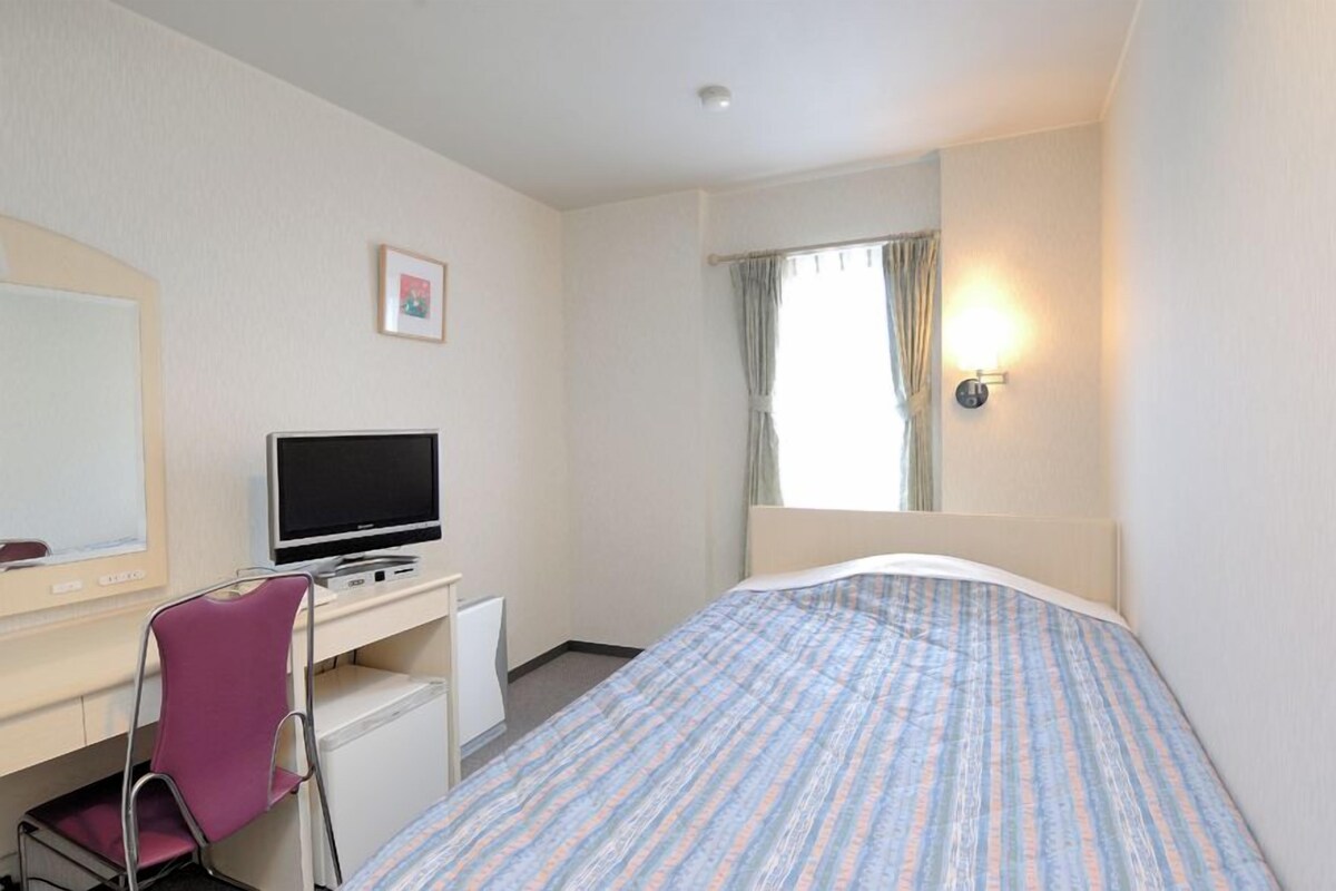A hotel room with a bed, a desk with a computer, a chair, a television, and a window with curtains.