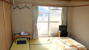 Une chambre d’hôtel avec un sol recouvert de tatamis, un lit avec une couvre-lit à motifs floraux, un bureau avec un téléphone et des papiers, un téléviseur posé sur un meuble et une porte coulissante donnant sur l’extérieur.