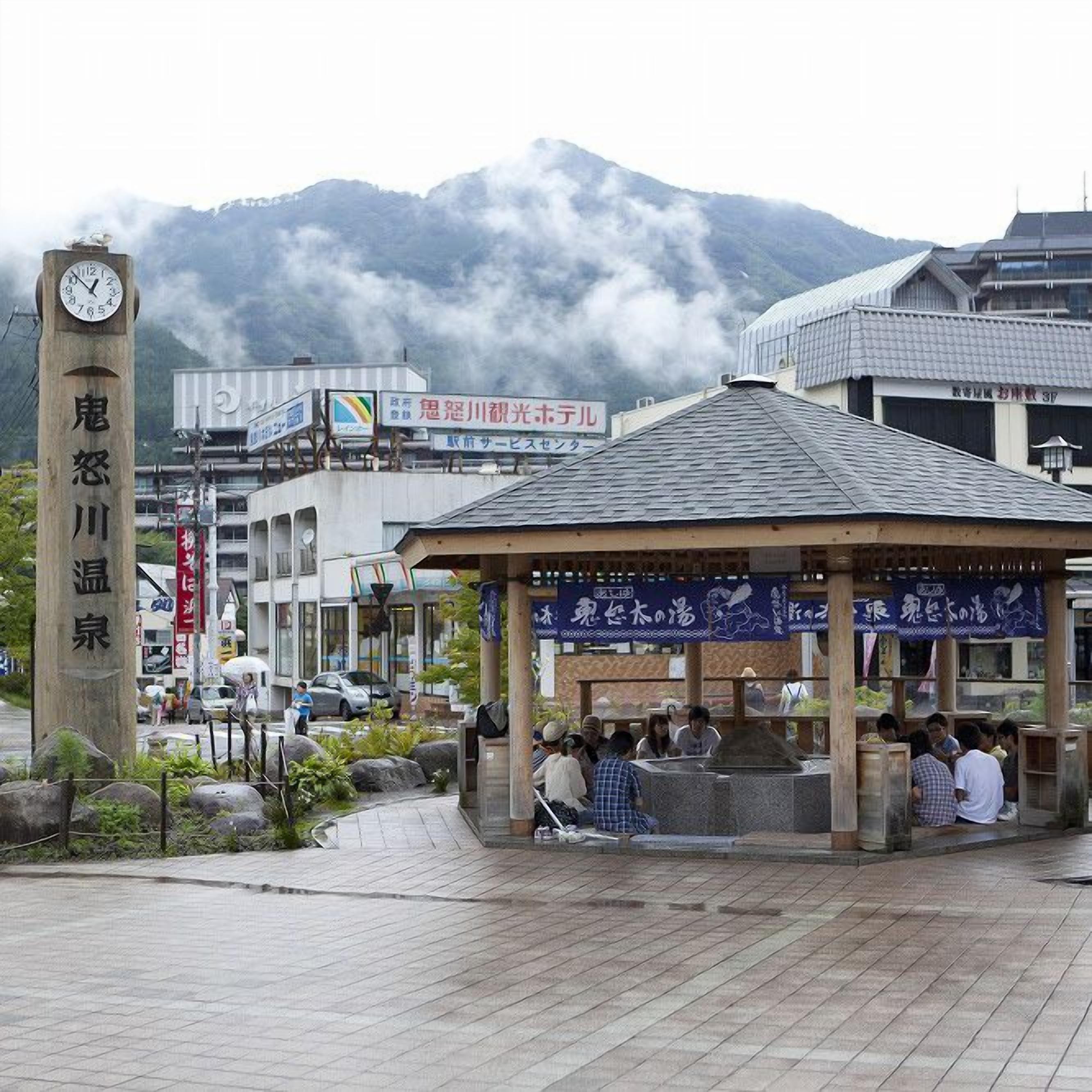 A public square with a clock tower, a covered seating area, and a mountain backdrop.