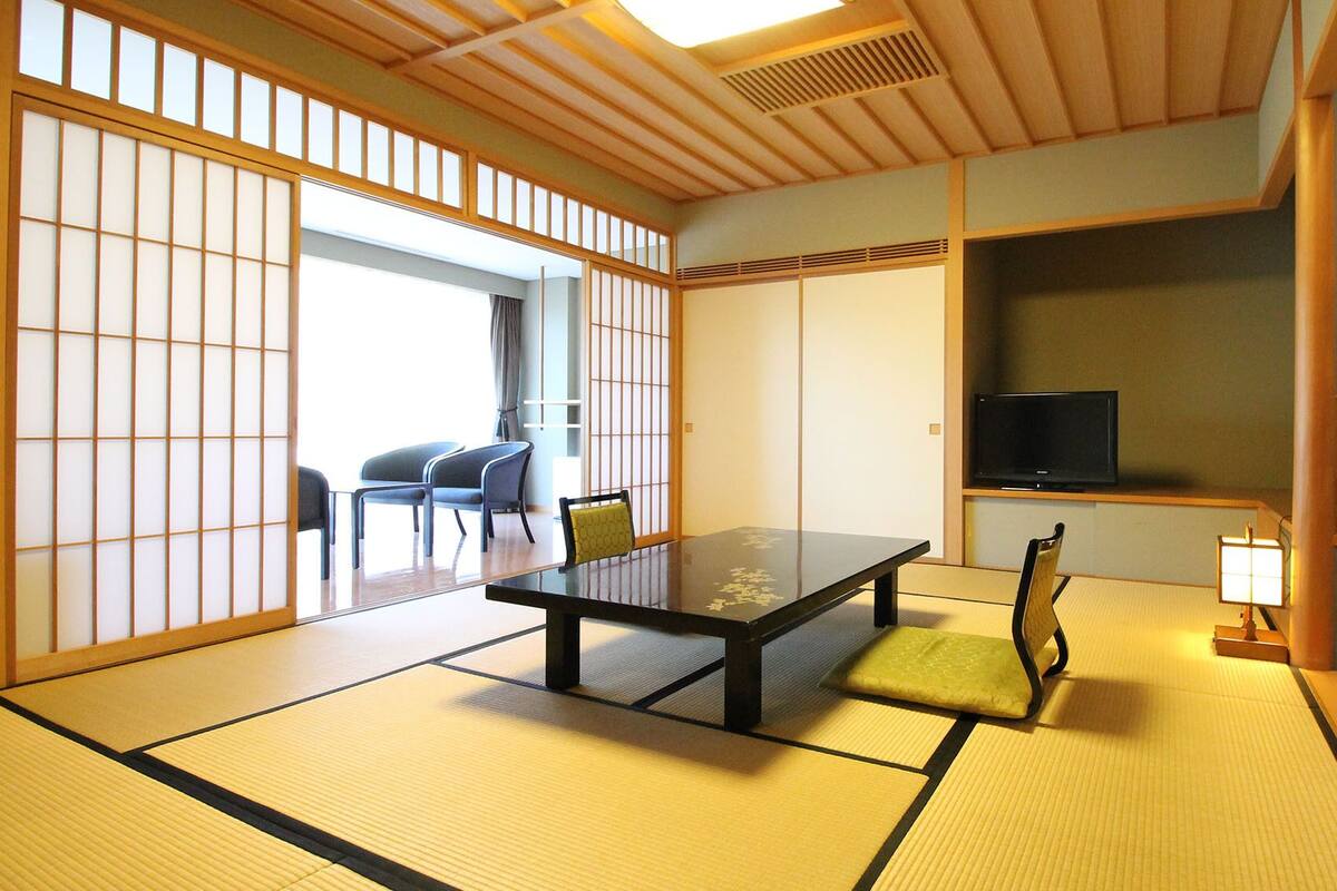 Een kamer met tatami matten, een lage tafel, stoelen, een televisietoestel en een schuifdeur.