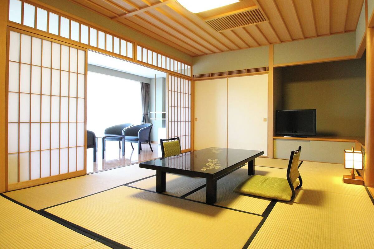 Een kamer met tatami matten, een lage tafel, stoelen, een televisie en een schuifdeur naar een andere kamer.