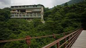 Un immeuble à plusieurs étages doté de balcons, entouré d’une forêt dense et surplombant un pont en bois.