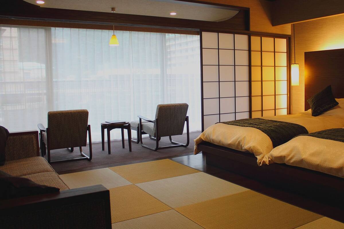 Hotelkamer met een groot bed, twee stoelen, een tafeltje en uitzicht op de stad door grote ramen.