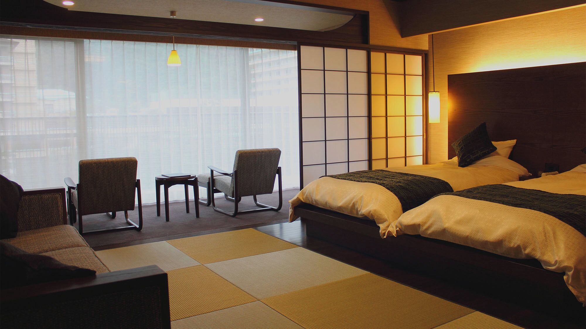 A hotel room with a bed, two chairs, a small table, and a view through large windows.