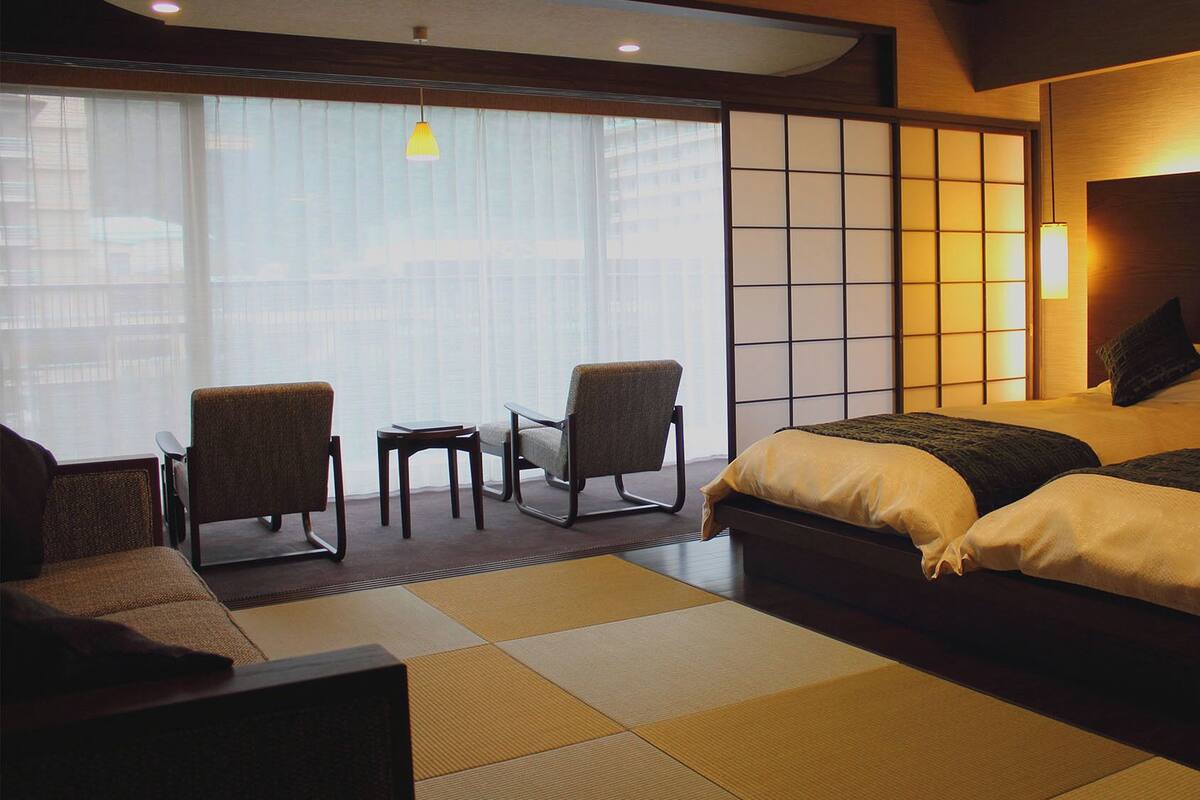 Een hotelkamer met een bed, twee stoelen, een tafeltje en uitzicht door grote ramen.