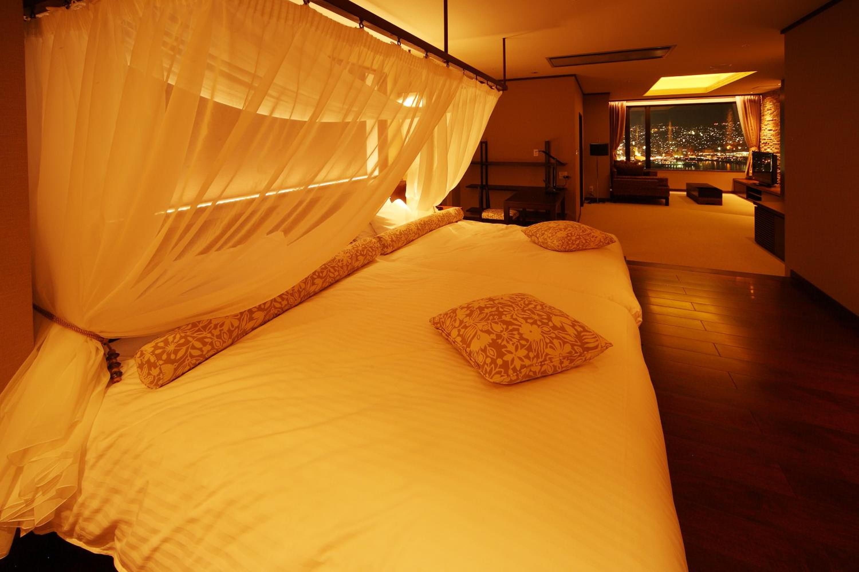 A bedroom with a canopy bed, a view of the city at night, and a seating area with a TV.