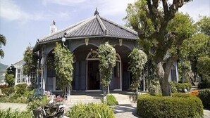 Une maison traditionnelle avec un toit en tuiles, une entrée centrale et un jardin bien entretenu, agrémenté d’un grand arbre et de plusieurs arbustes.