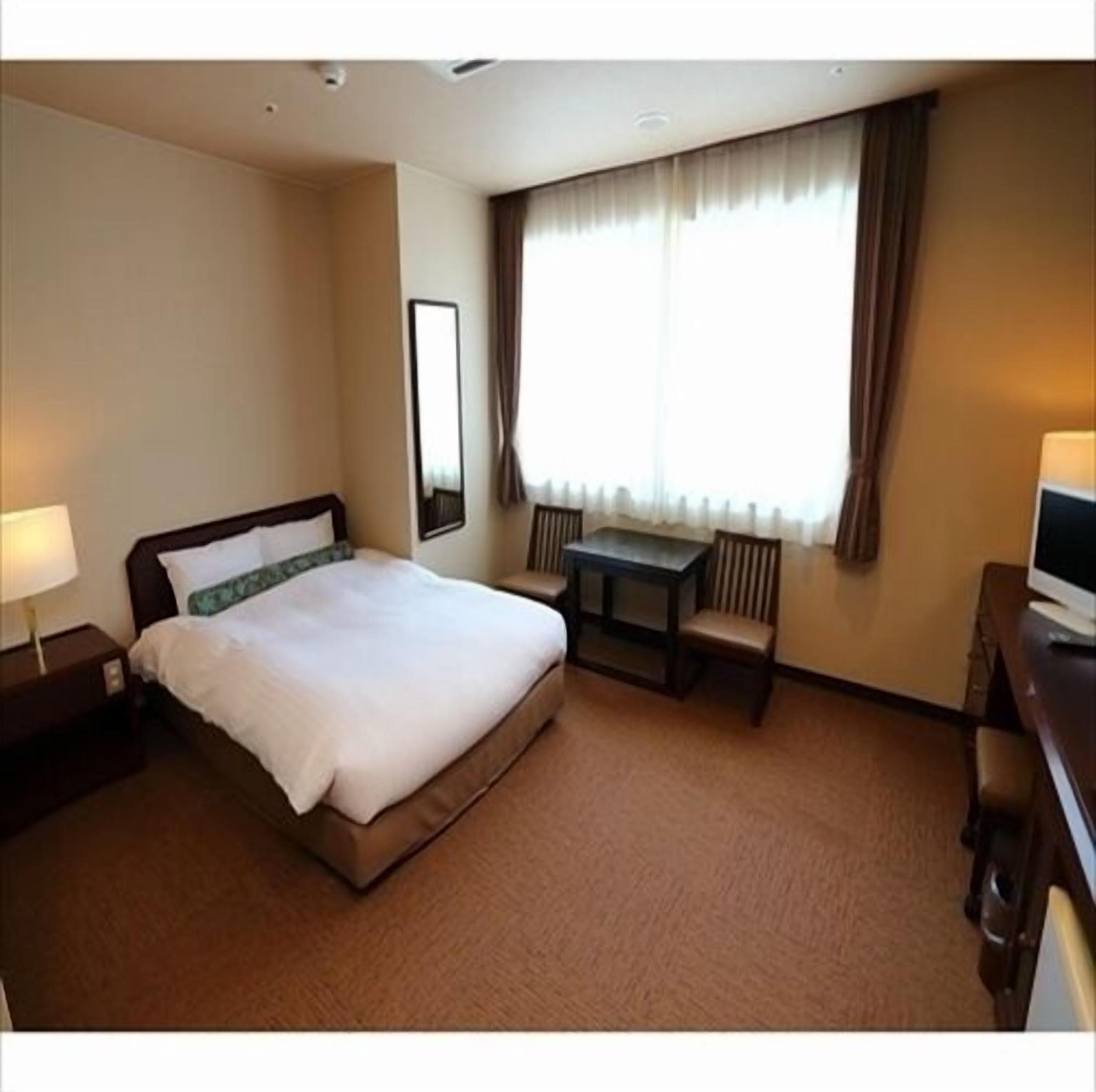 Habitación de hotel con cama, escritorio, silla, televisión y ventana con cortinas.