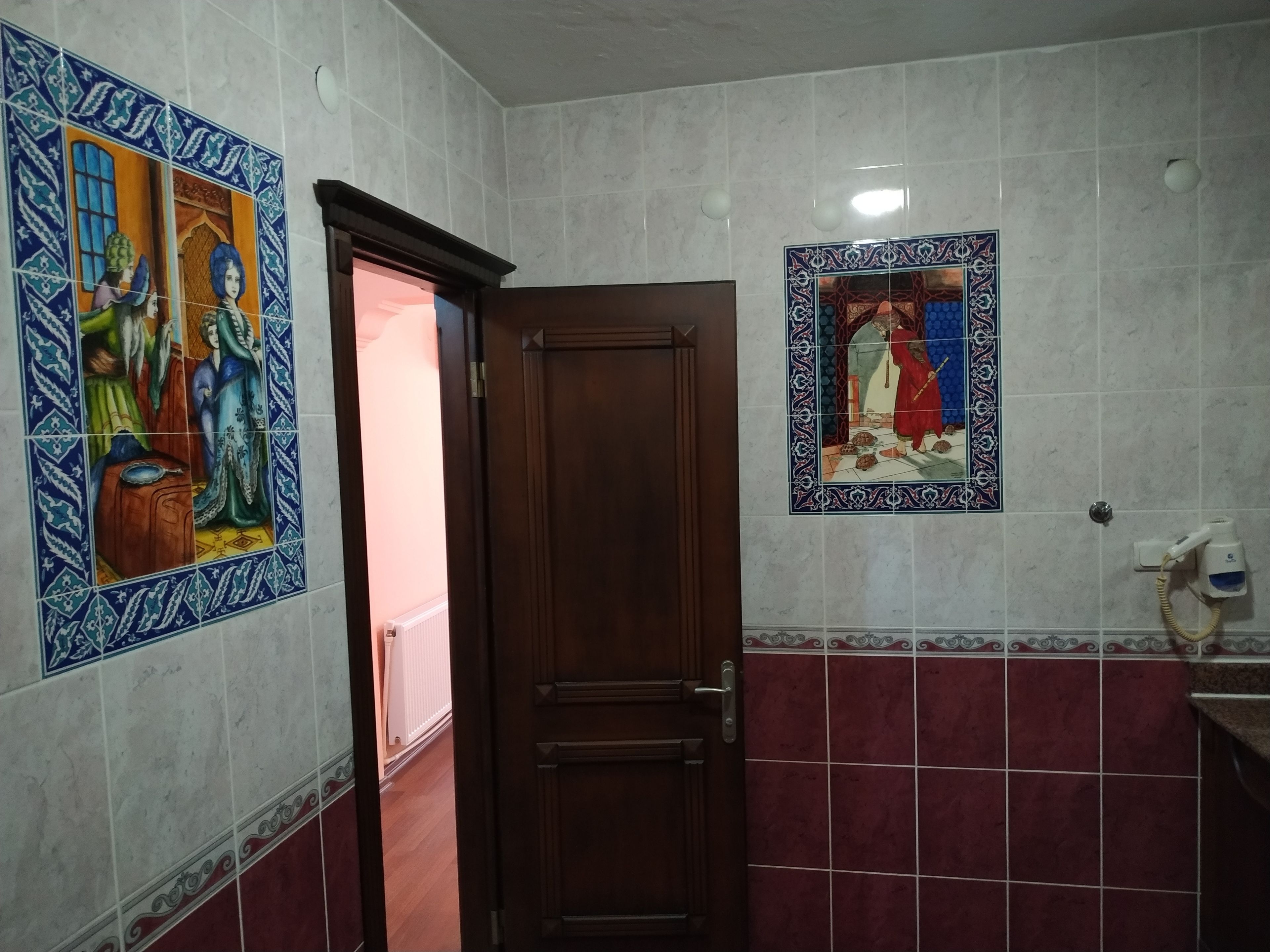 A bathroom with a wooden door, a tiled wall with a decorative tile featuring a person, and a hand dryer on the wall.
