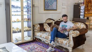 Un homme est assis sur un canapé à motifs dans une pièce où l’on aperçoit une vitrine contenant des chaussures, un tapis à motifs et une décoration murale.