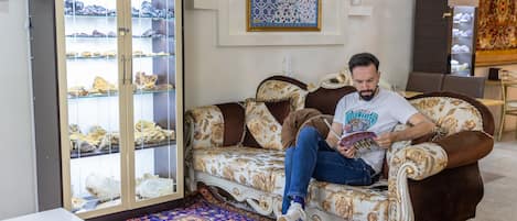 Un hombre sentado en un sofá estampado en una habitación con una vitrina de zapatos, un tapete decorativo y un adorno en la pared.