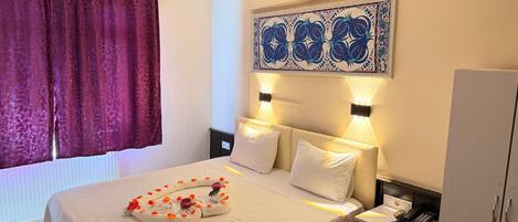 Quarto de hotel com cama, duas luminárias de cabeceira, um ornamento na parede, um guarda-roupa e uma janela com cortinas.