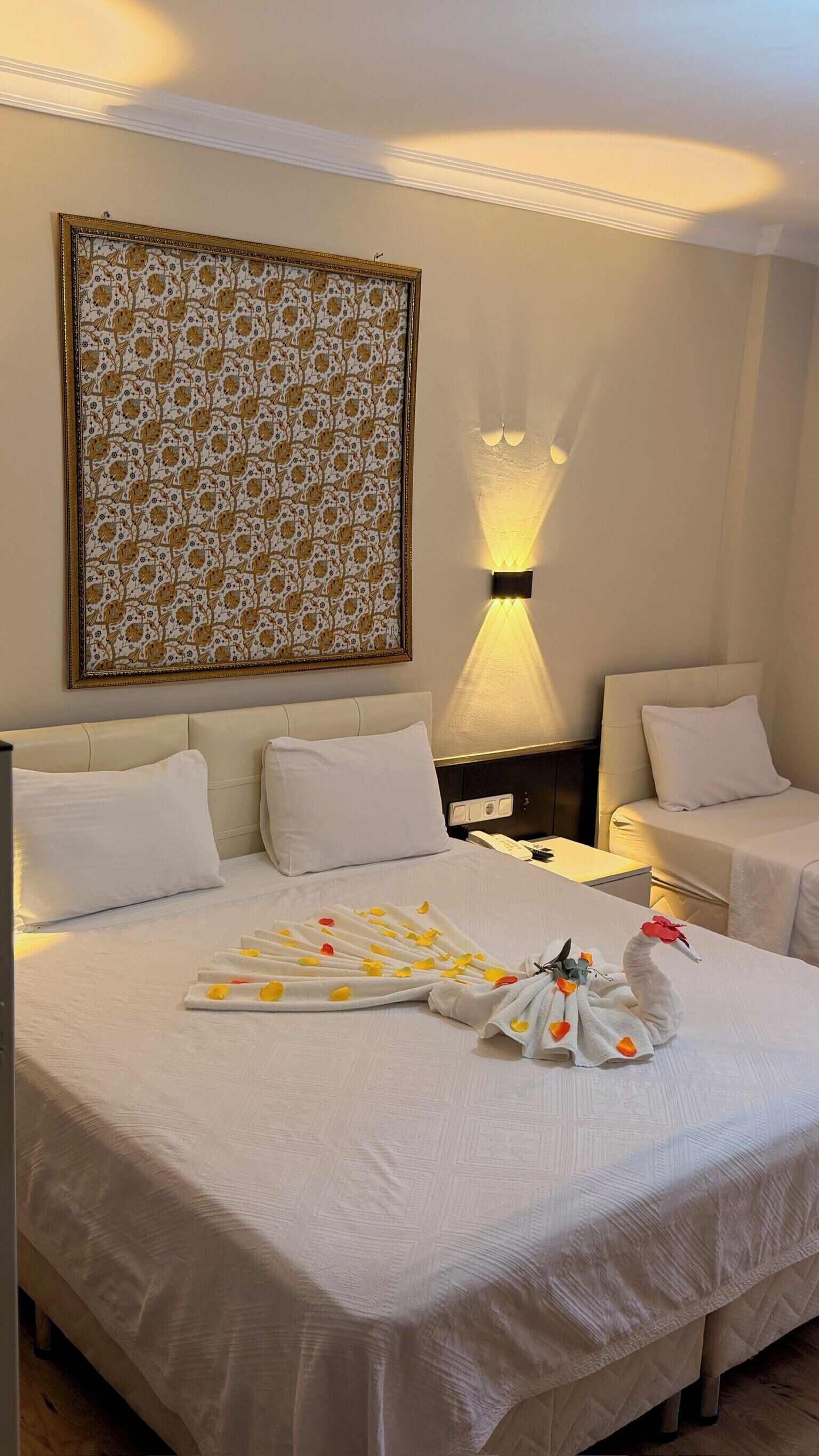 Una habitación de hotel con una cama grande, dos mesitas de noche, una lámpara de pared y una obra de arte floral enmarco.