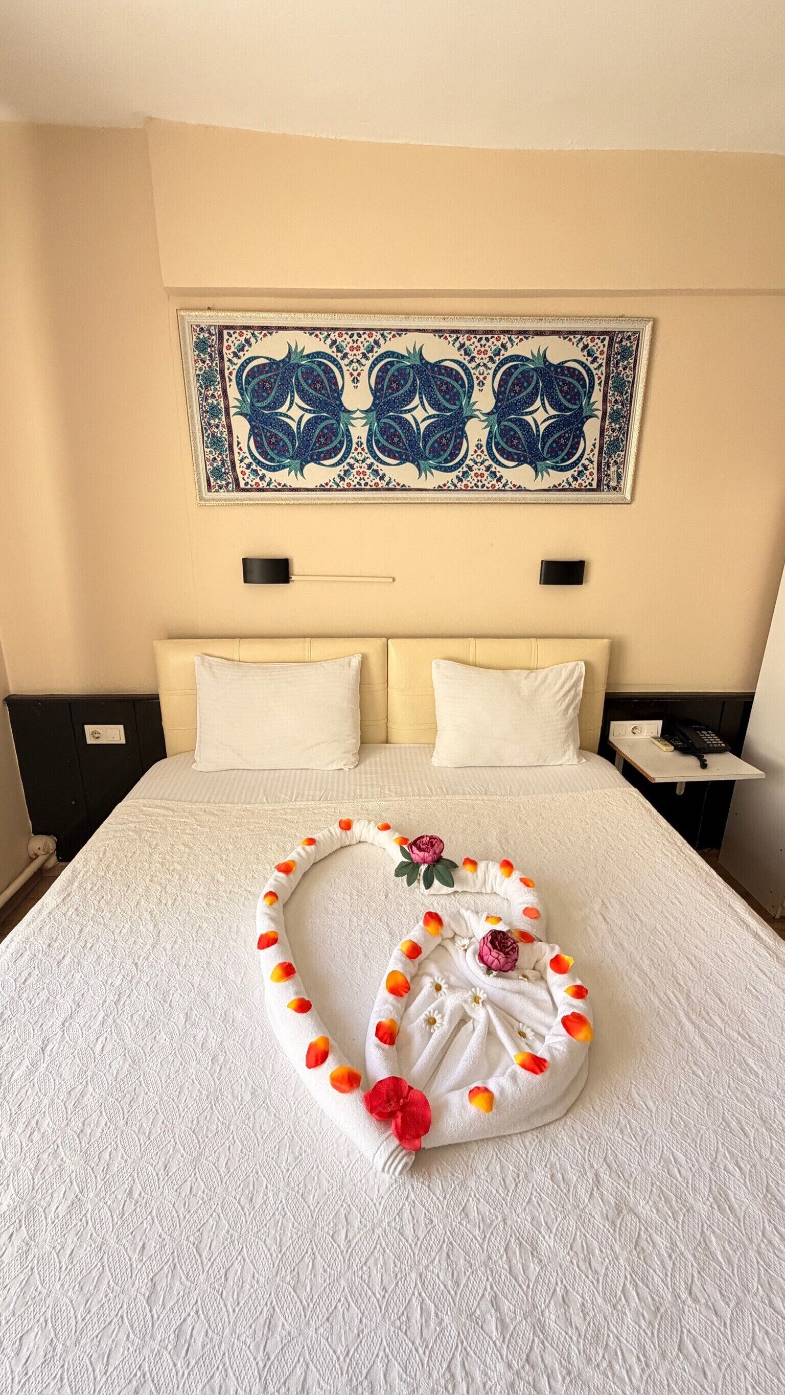 Une chambre d’hôtel avec un grand lit, une décoration en forme de cœur posée sur le lit, une tapisserie murale et une table de chevet avec un téléphone.