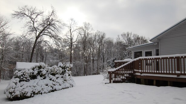 A snow-covered deck with a wooden railing, a snow-covered bush, and a house with a visible garage door. - Comfortable lake living, pool access, 3 miles to marina (Monticello)