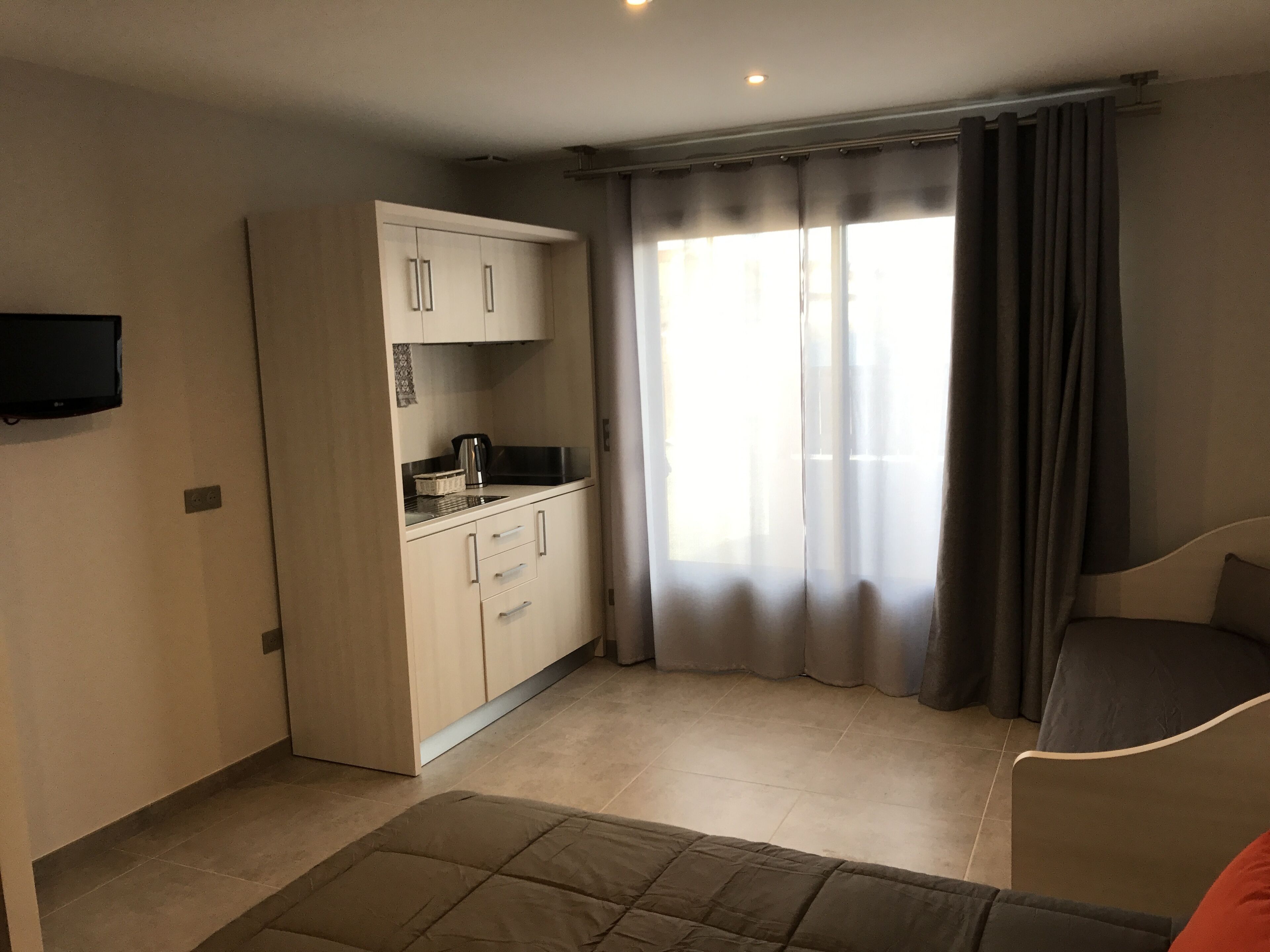Ein kompakter Wohnbereich mit Küchenzeile, einem wandmontierten Flachbildfernseher, einer Couch und einem großen Fenster mit Vorhängen.