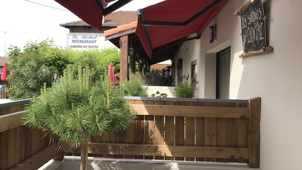 Ein Sitzbereich im Freien mit Holzbänken und roten Sonnenschirmen, ein Schild für das Restaurant 'Les Studios du Centre' und Blick ins grüne Umland.