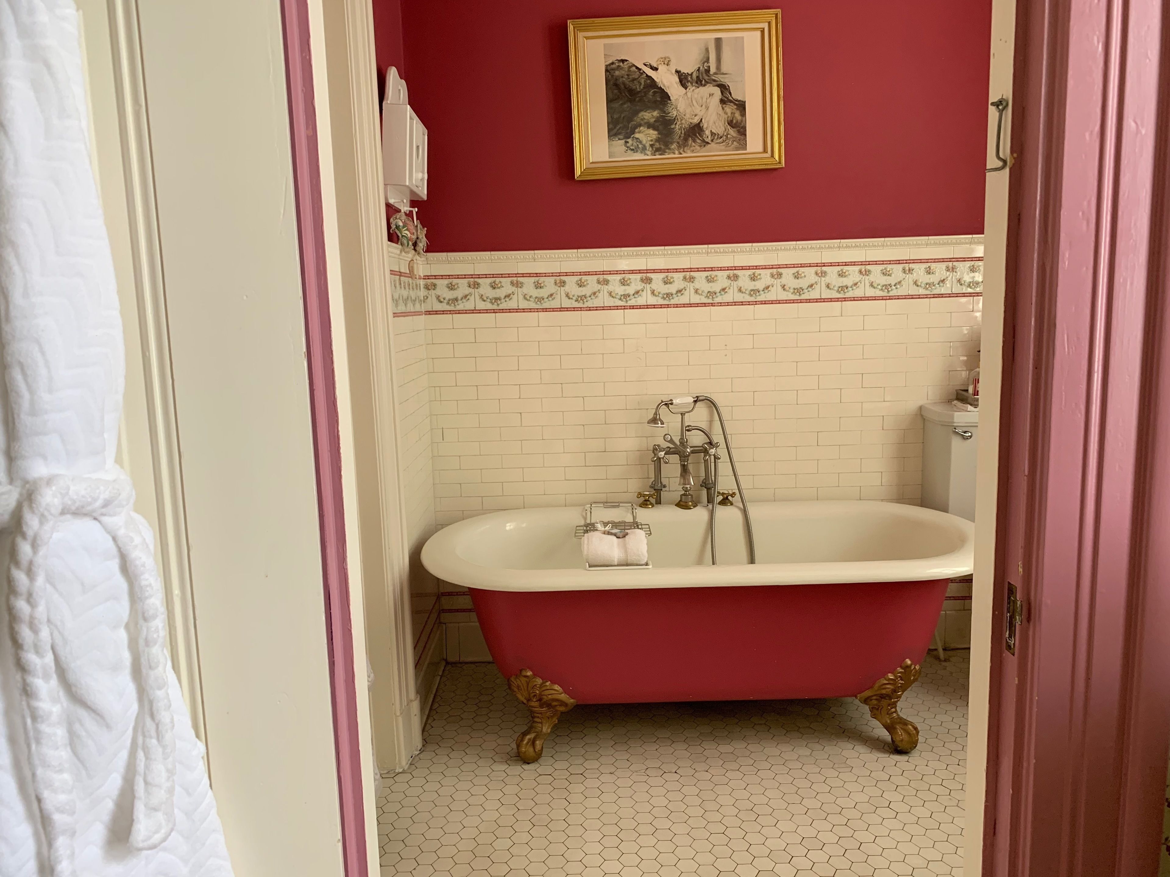 A bathroom with a clawfoot bathtub, a white toilet, and a red wall with a framed picture.