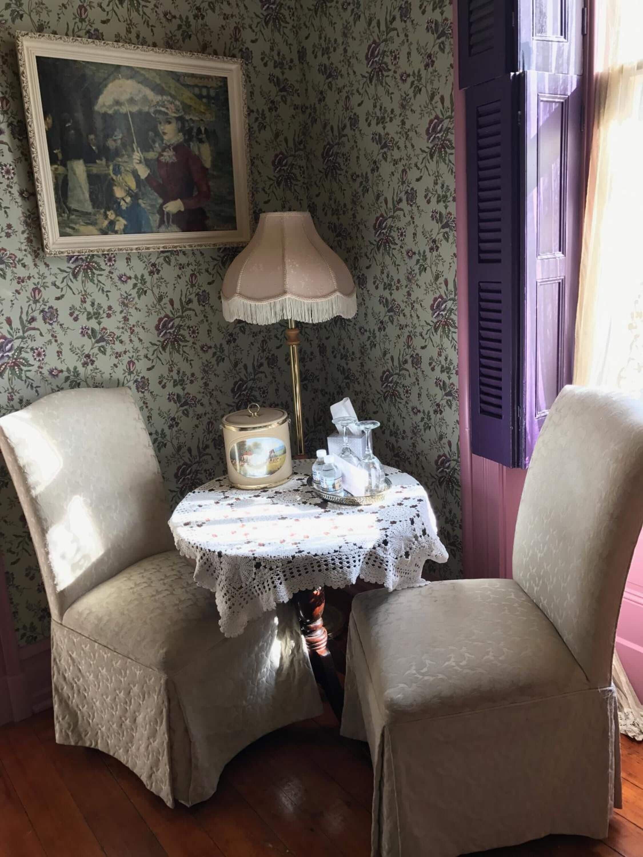 A corner room with floral wallpaper, a table with a lace tablecloth, a lamp, and a painting on the wall.