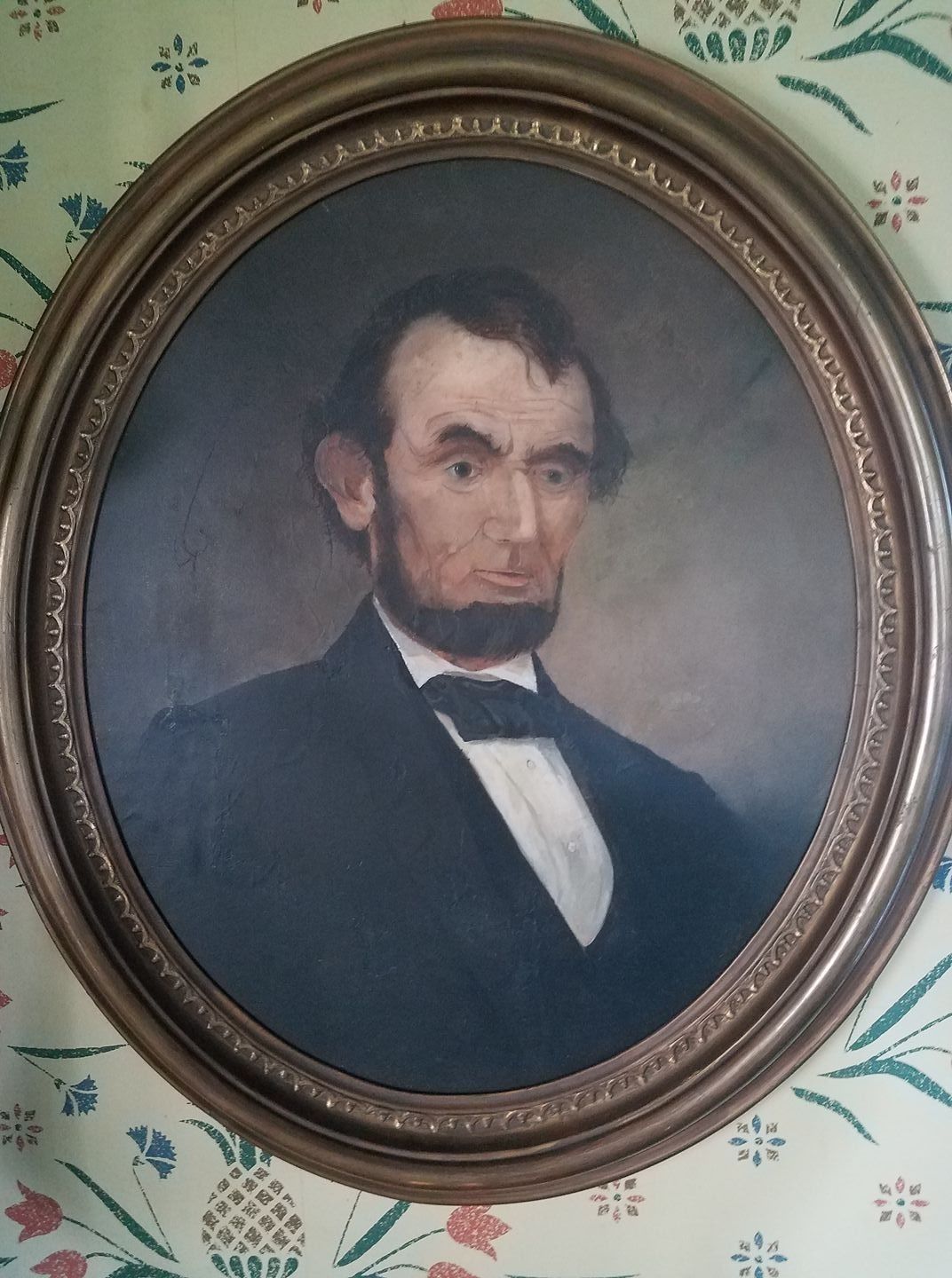 A portrait framed in a decorative border, mounted on a wall with a floral patterned wallpaper.