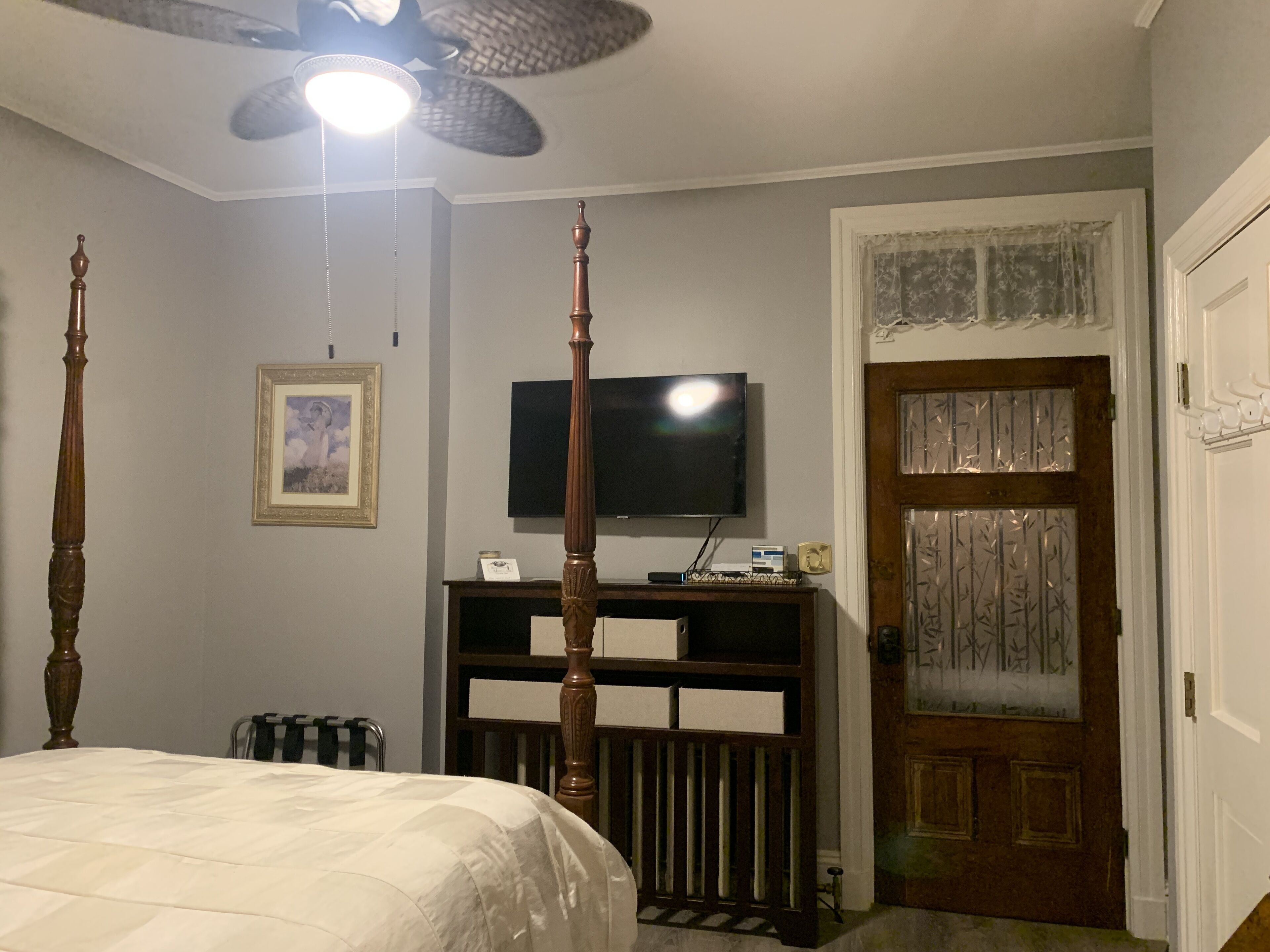 Un dormitorio con cama con dosel, ventilador de techo, televisor sobre un soporte de madera y puerta con paneles de vidrio decorativos.