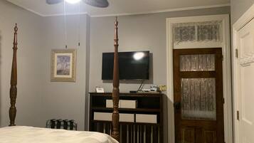 Un dormitorio con cama con dosel, ventilador de techo, televisor sobre un soporte de madera y puerta con paneles de vidrio decorativos.