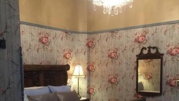 A bedroom with a wooden bed, floral wallpaper, a chandelier, and a mirror. - Susquehanna Manor Inn (Marietta)