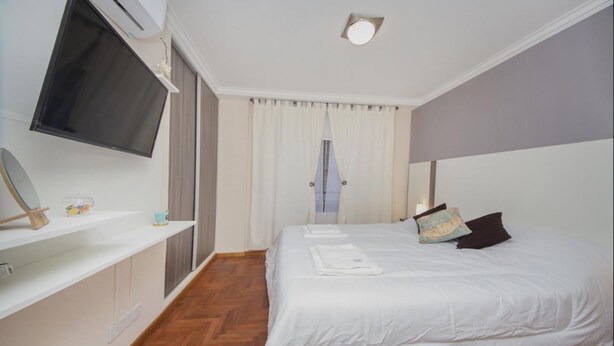 A hotel room with a bed, a TV mounted on the wall, a window with curtains, and a wooden floor. - San Juan Flat 33W (Rio Cuarto)