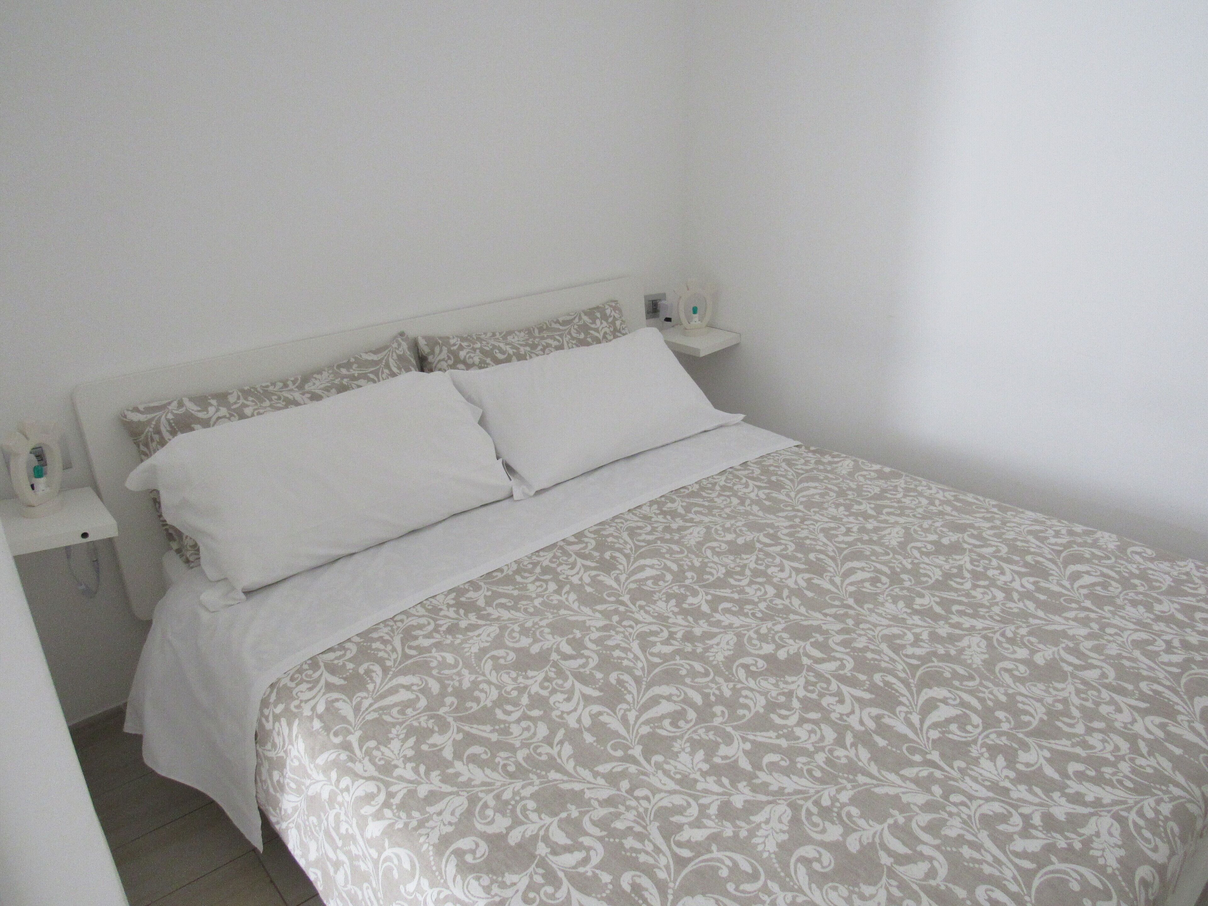 Un lit bien fait, avec une couvre-lit à motifs, des oreillers blancs et une table de chevet sur laquelle se trouvent une lampe et une bouteille.