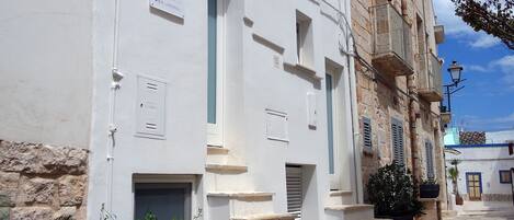Una calle estrecha con edificios blancos, algunos con ventanas y puertas visibles, y unas cuantas plantas en macetas.