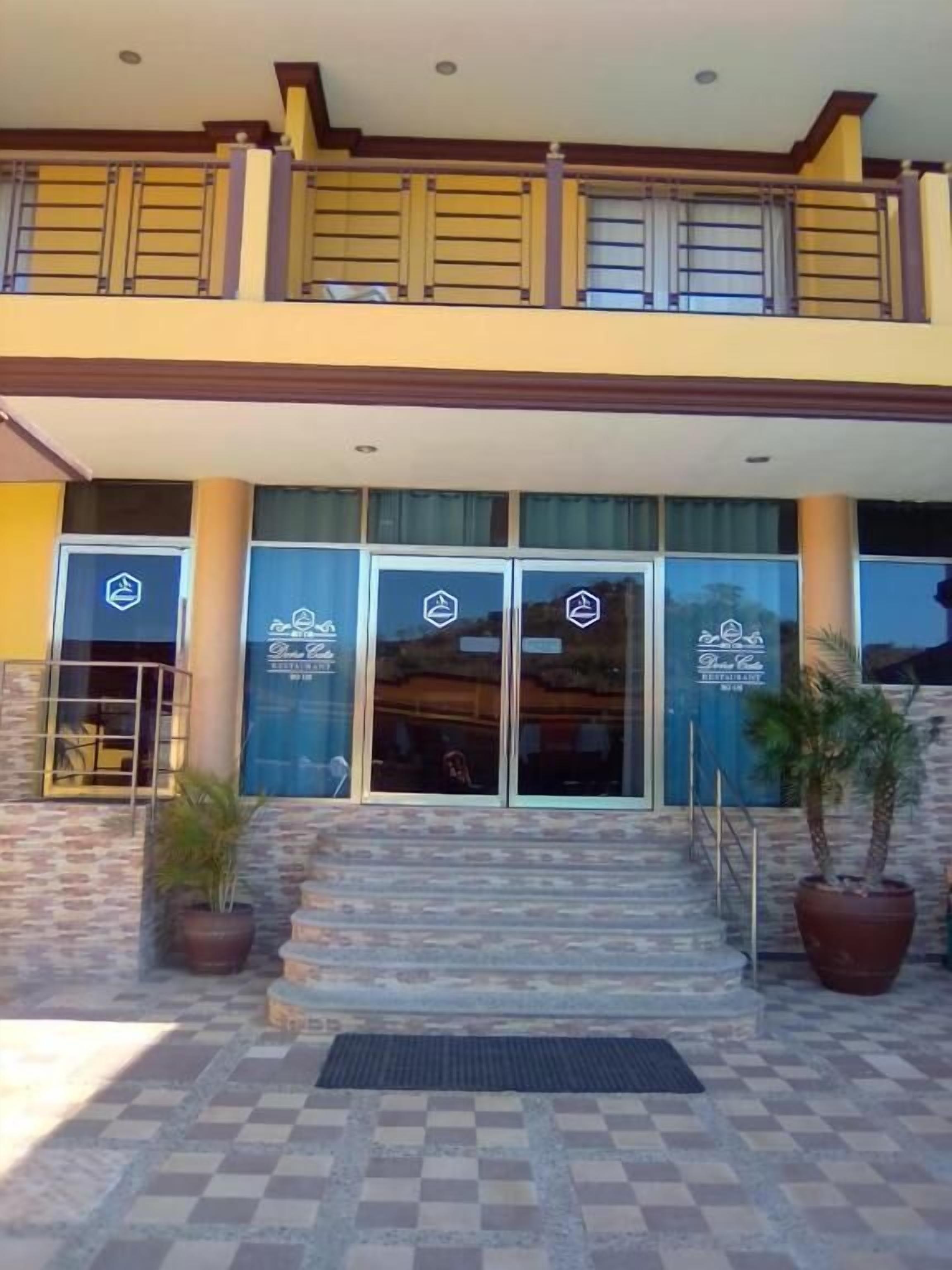A hotel entrance with a checkered tile floor, a set of stairs leading up to the doors, and two potted plants on either side.