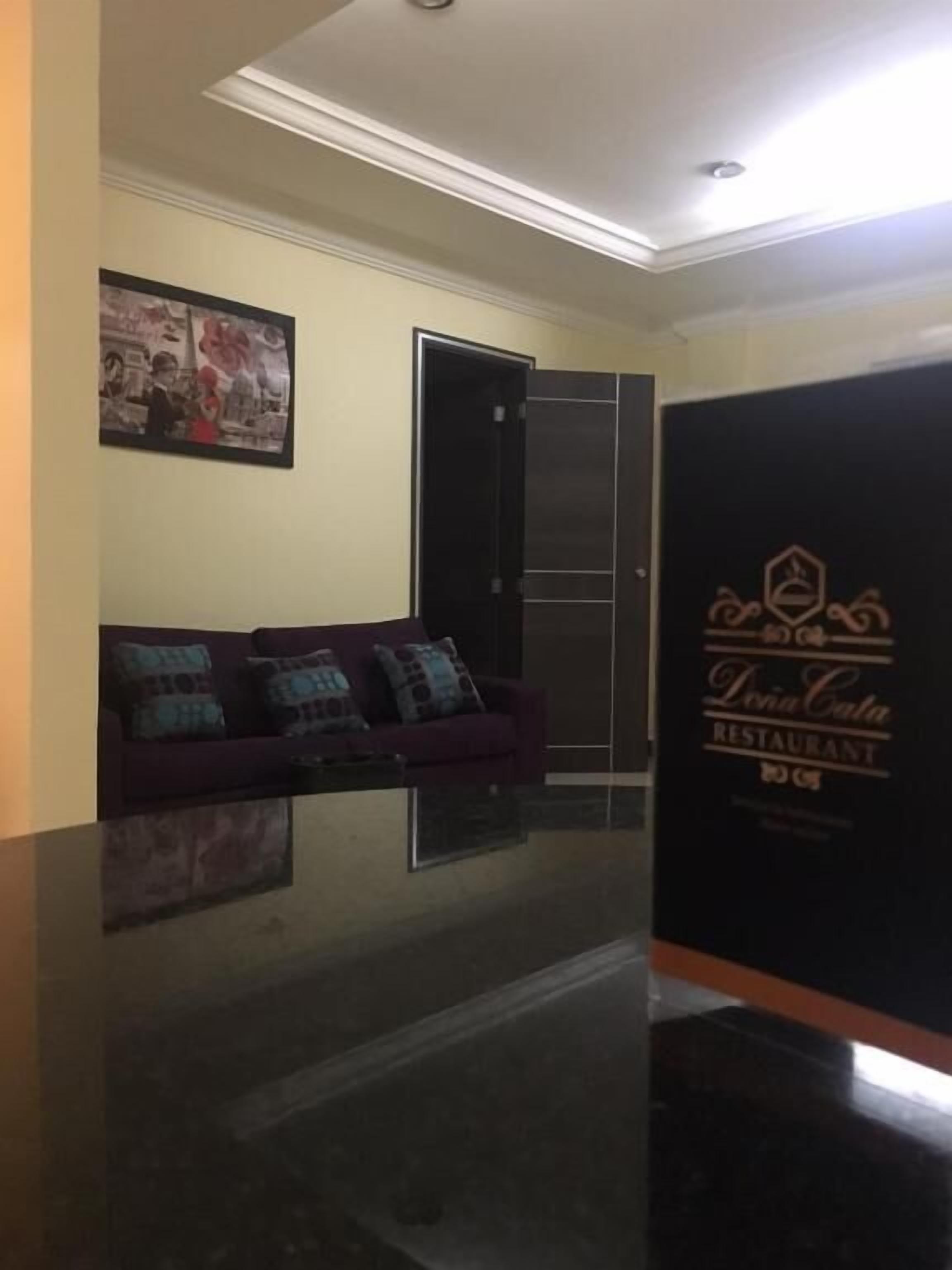 Un área de recepción con un mostrador oscuro, un sofá morado con cojines estampados, un cuadro en la pared y una puerta que da al restaurante.