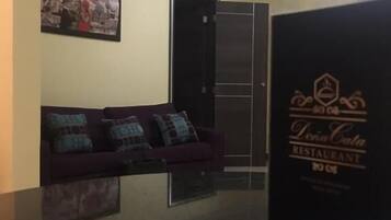 Un área de recepción con un mostrador oscuro, un sofá morado con cojines estampados, un cuadro en la pared y una puerta que da al restaurante.
