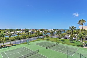 Cancha de tenis con red, rodeada por una valla, y con vista a un cuerpo de agua con edificios y palmeras al fondo.