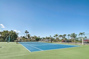 Una cancha de tenis al aire libre con red, rodeada de palmeras y un cielo despejado.