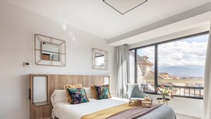 Een moderne hotelkamer met een groot bed, een houten hoofdbord, twee spiegels aan de muur, een balkon met uitzicht op de stad, en een tafeltje met een bloevas.