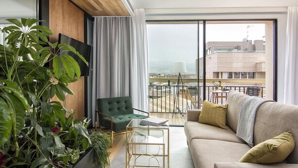 Een moderne woonkamer met een bank, een groene fauteuil, een glazen salontafel en een groot raam met uitzicht op gebouwen en een wateroppervlak.