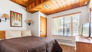 Un dormitorio con una cama grande, techo de madera, dos lámparas de pared, una ventana con persianas y un aparador de madera.
