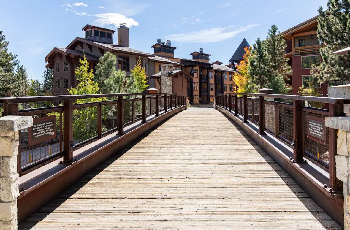 Un puente de madera que conduce a un albergue con varias construcciones, rodeado de árboles.