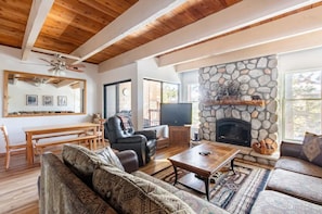 A cozy living room with a stone fireplace, wooden ceiling beams, and a large window.