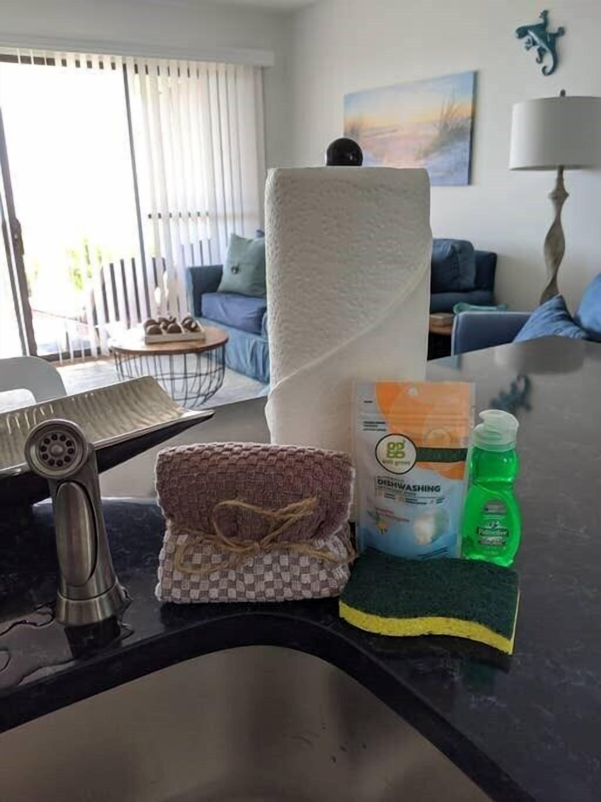 A kitchen counter with a paper towel roll, a woven cloth, a dishwashing liquid bottle, and a sponge.